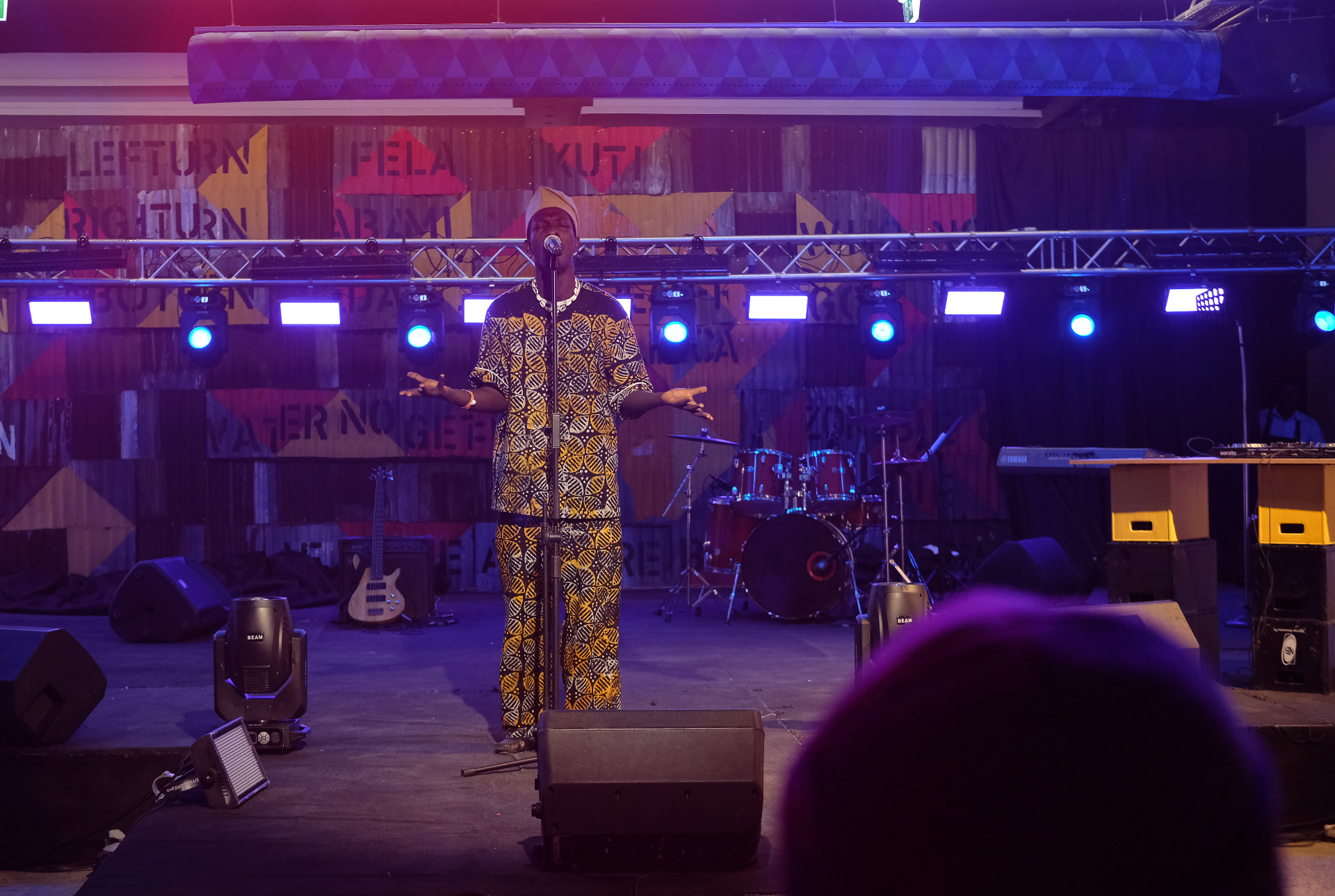 Performer in patterned dress speaking at center stage beneath blue and violet lights at Afrobeat Rebellion