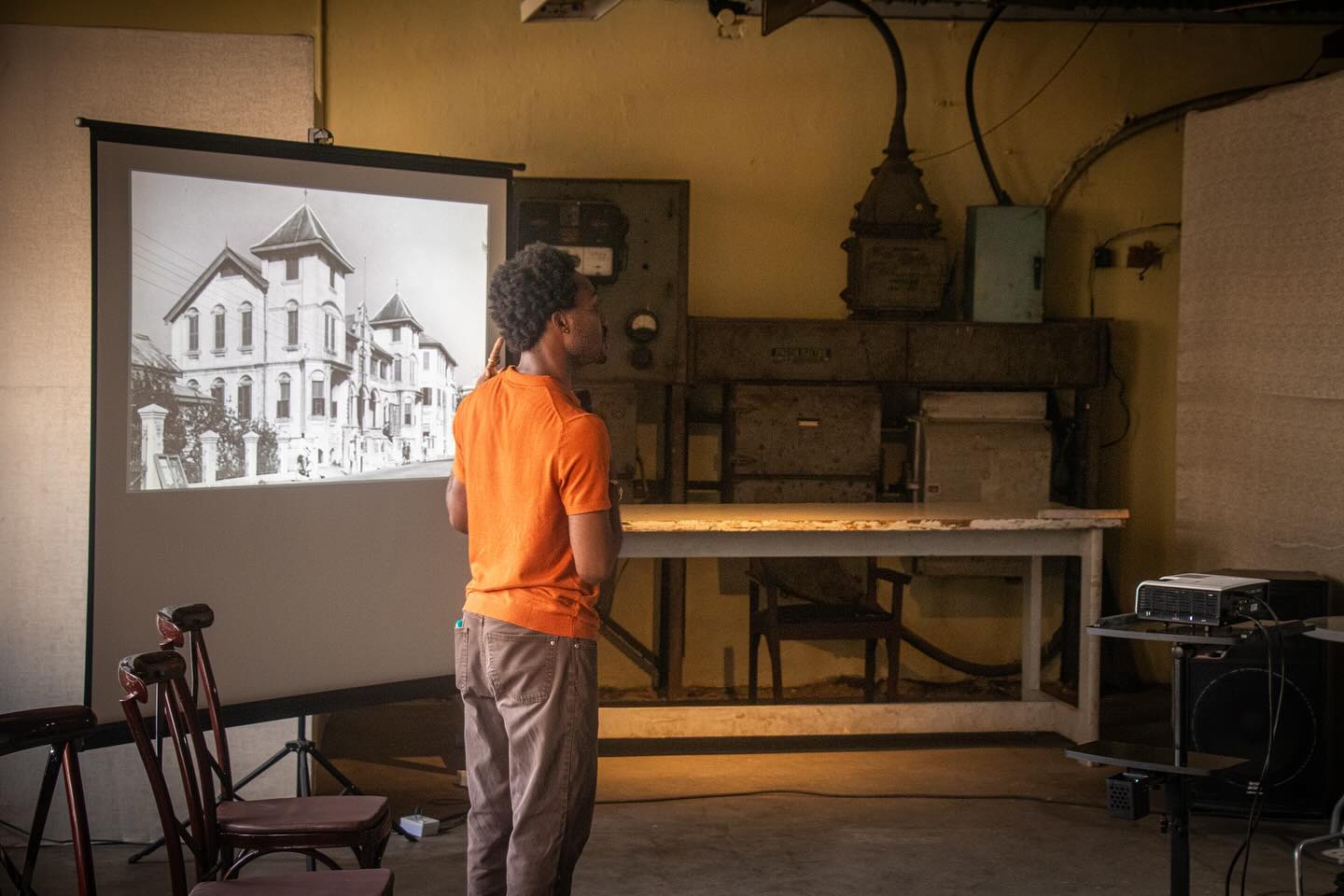Speaker in orange presenting a black-and-white archival image of colonial Lagos architecture against the raw machinery of the old Government Printing Press