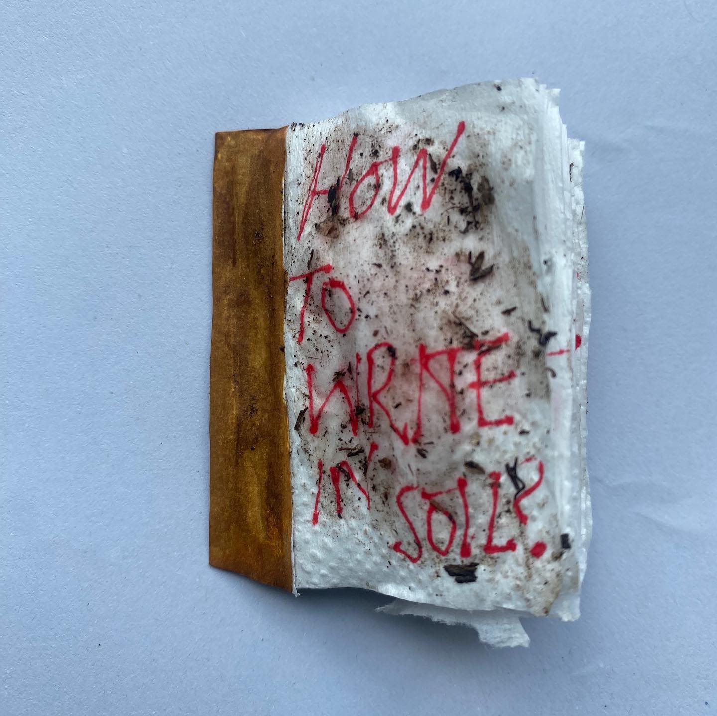 Small hand-bound zine titled 'How to Write in Soil' with red hand-lettered text and earth pressed into the pages
