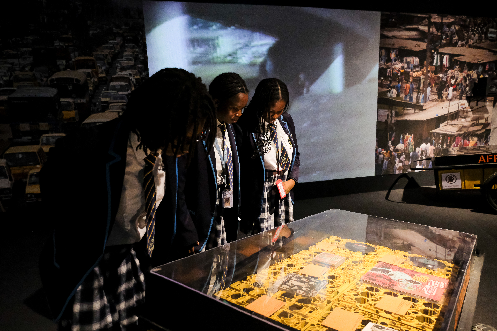 Schoolgirls in uniform examining the yellow vinyl record vitrine beside Lagos street projections at Afrobeat Rebellion